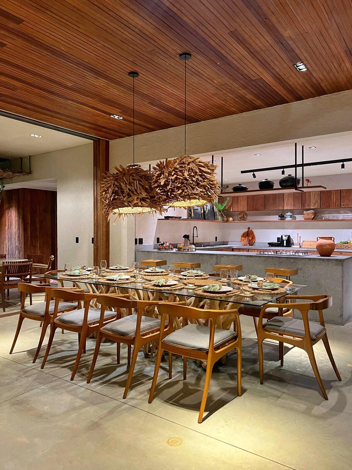 Cozinha ao fundo da mesa de jantar, sob forro de madeira e lustres de palha