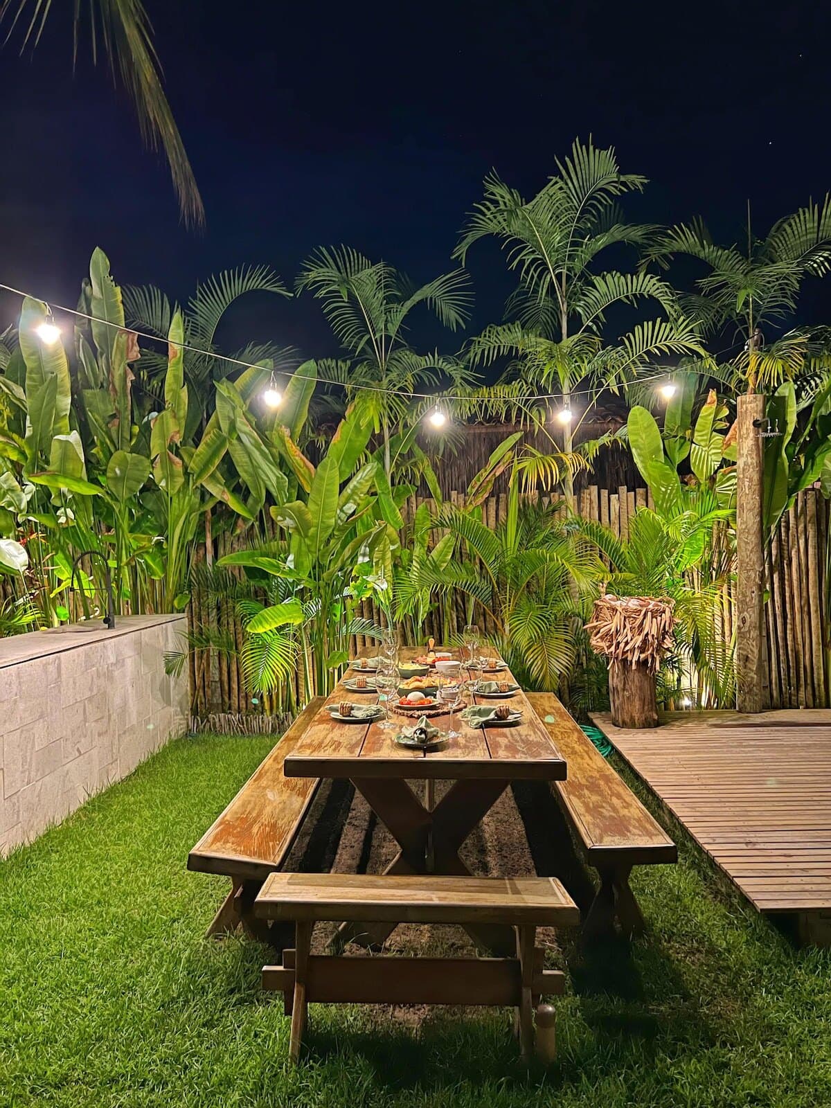 Jardim à noite com lâmpadas penduradas, mesa de madeira com bancos no gramado e bambu ao fundo
