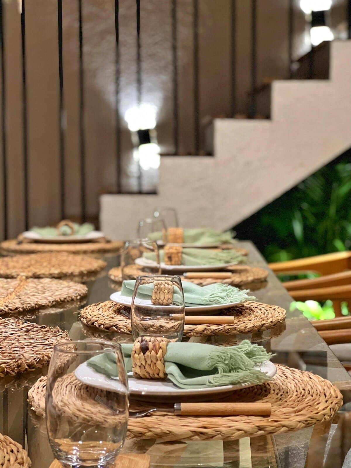 Detalhe da mesa posta com sousplat de fibra trançada, guardanapo verde-sálvia e a escada ao fundo