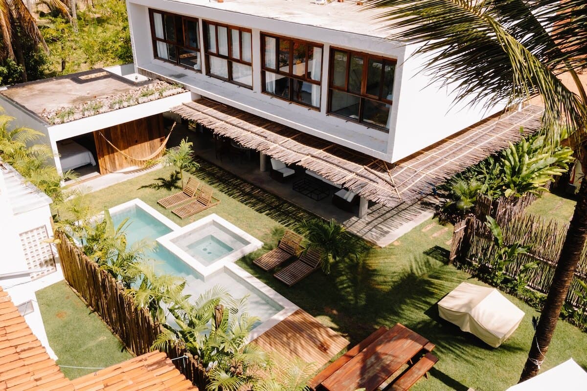 Vista de cima da Casa Tropi, com o sobrado branco de janelas de madeira, a piscina com jacuzzi, o gramado, o deck e as palmeiras
