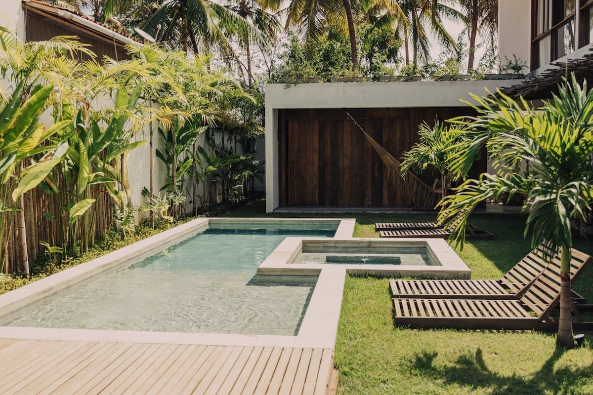 Piscina e jacuzzi de borda de pedra clara, com deck de madeira, espreguiçadeiras de ripas, bambu e palmeiras