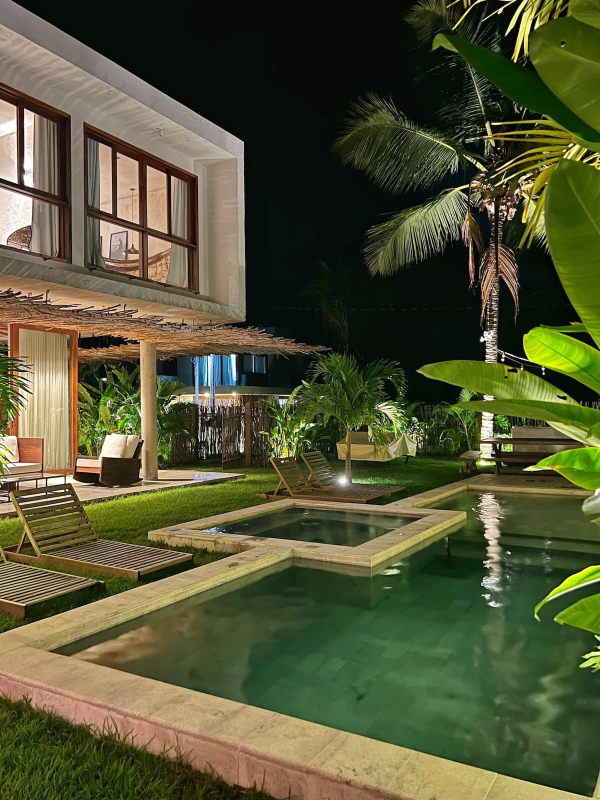 Casa Tropi à noite, com a piscina e a jacuzzi iluminadas, as janelas acesas do sobrado e palmeiras no jardim