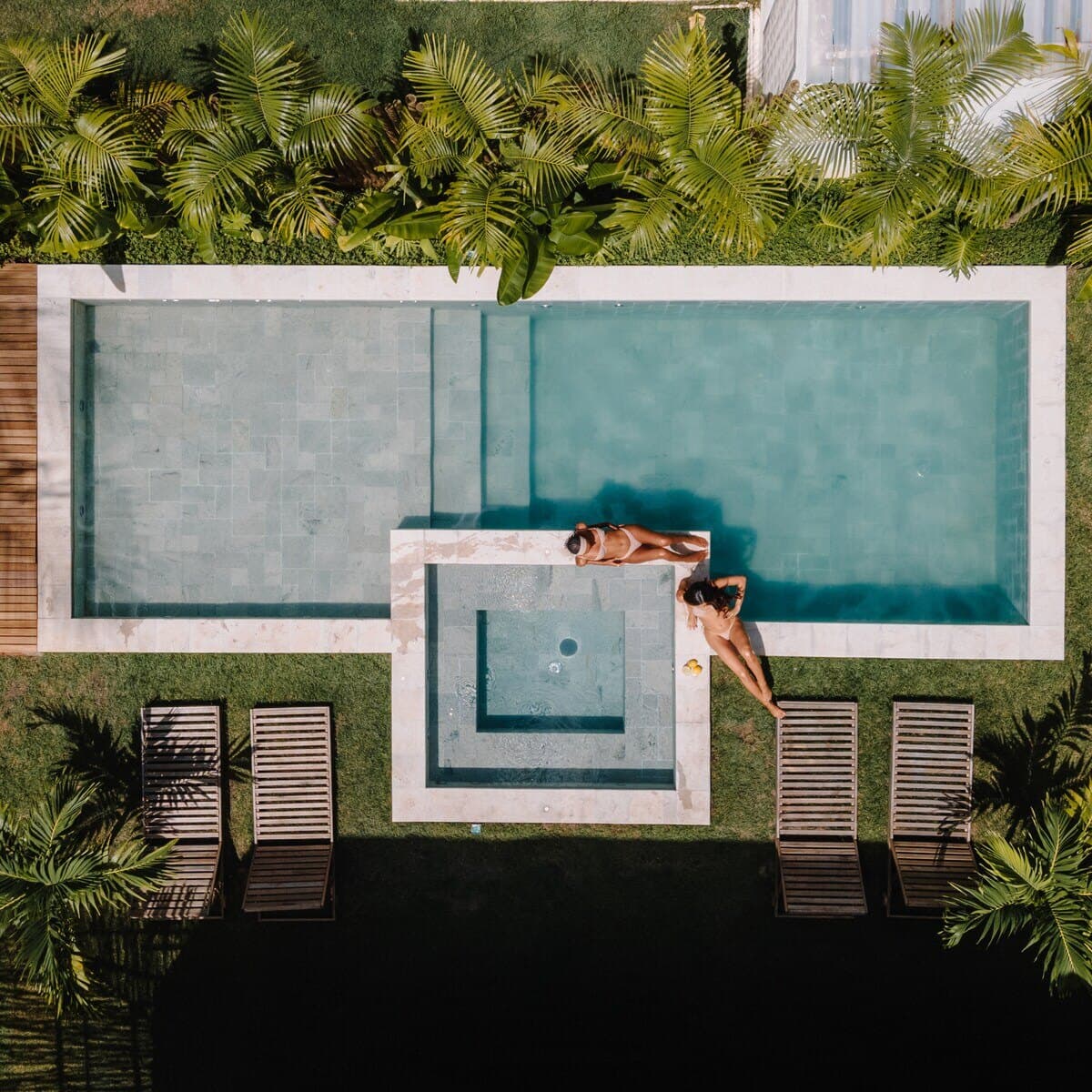 Piscina comprida vista de cima, com jacuzzi quadrada de borda clara, espreguiçadeiras de ripas, gramado e palmeiras ao redor