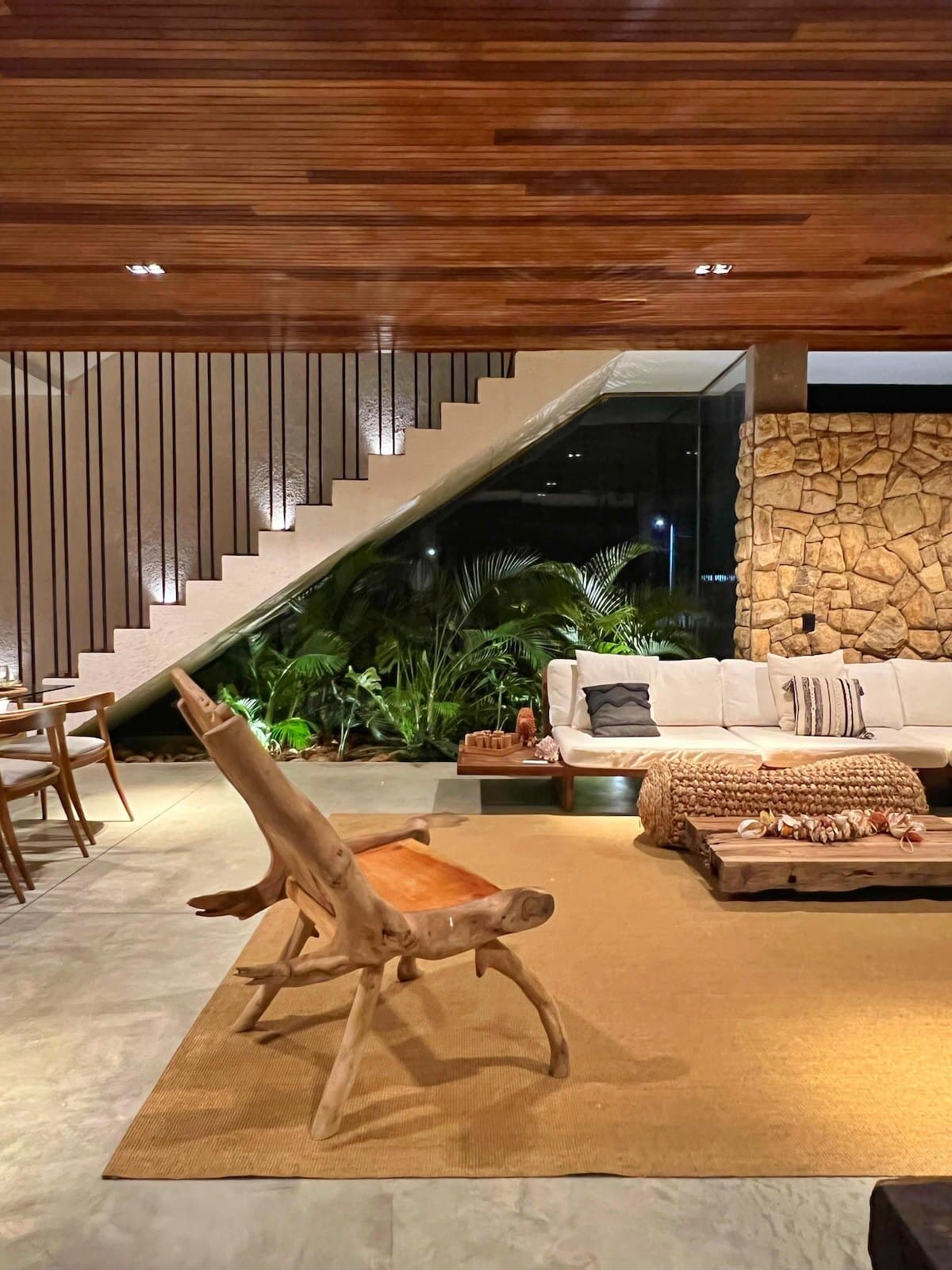 Escada flutuando sobre plantas dentro da sala, com forro de madeira, parede de pedra e sofá ao fundo